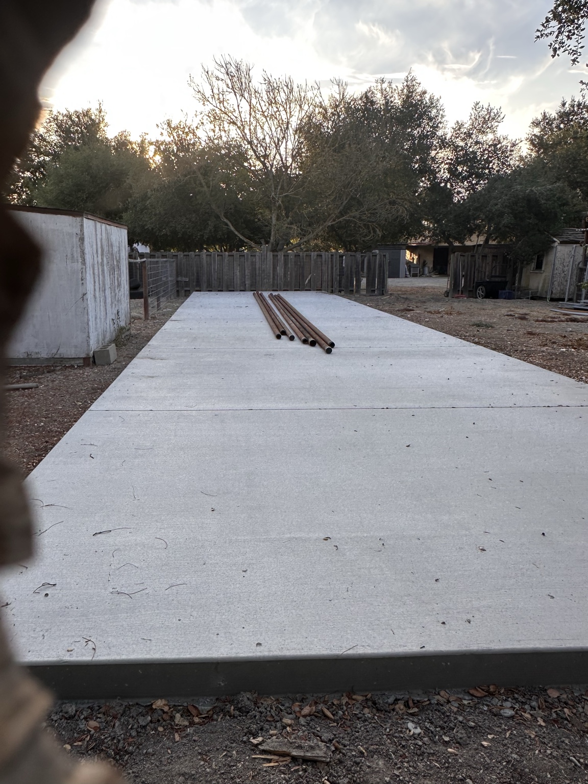 Large concrete slab with several steel pipes resting on it beside fencing and outbuildings
